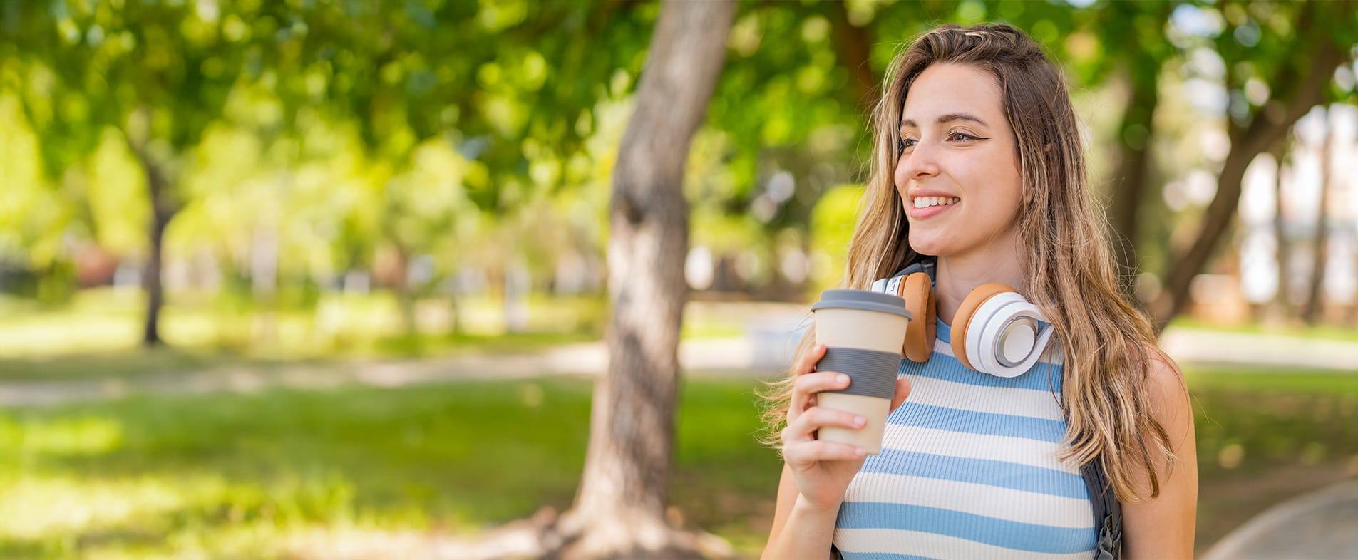 Ondernemer met koptelefoon om haar nek en koffie in haar hand, lachend in een park